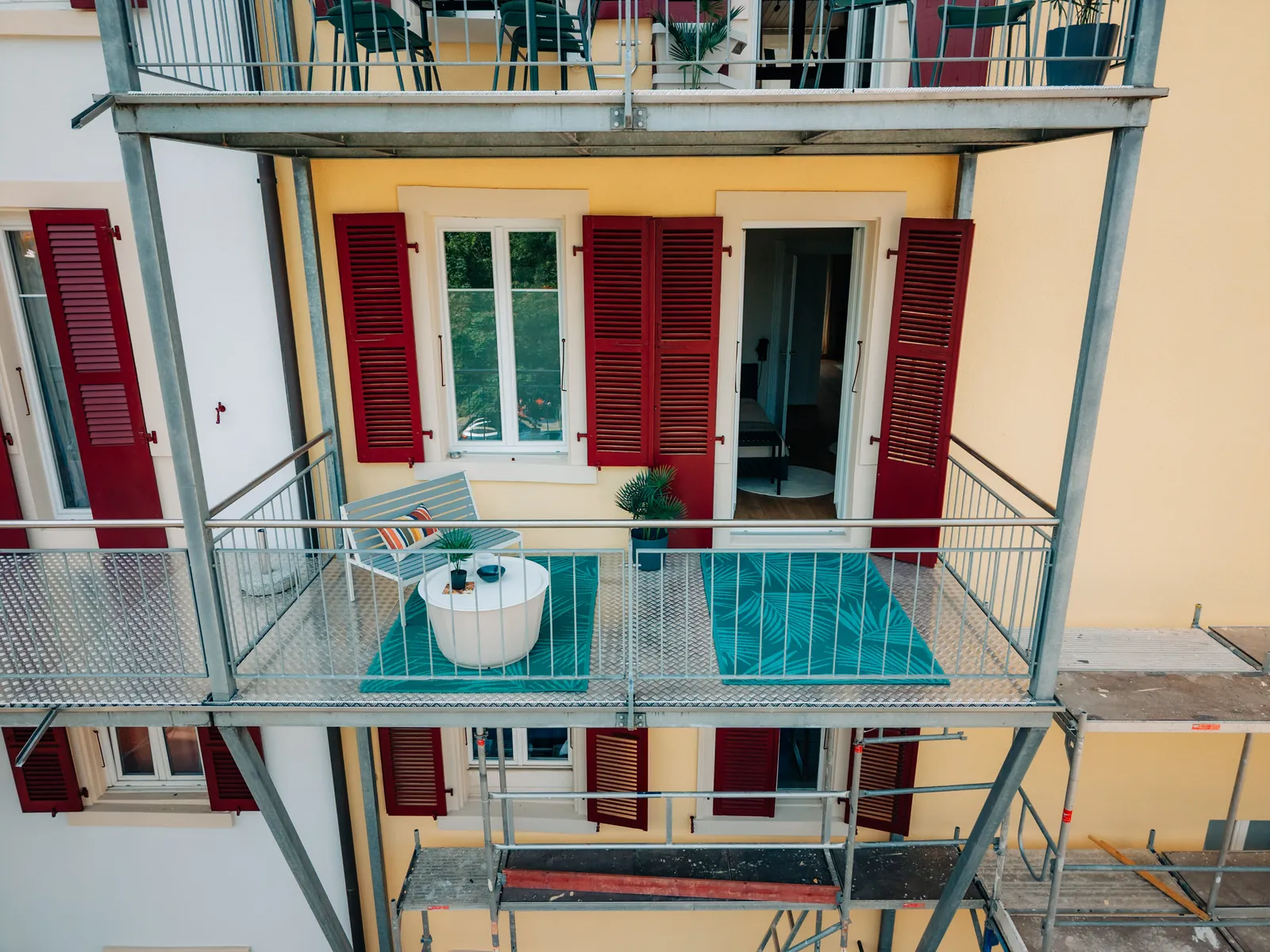 Möblierter Balkon mit Bank, Beistelltisch und Teppich vor der gelben Fassade mit weinroten Fensterläden